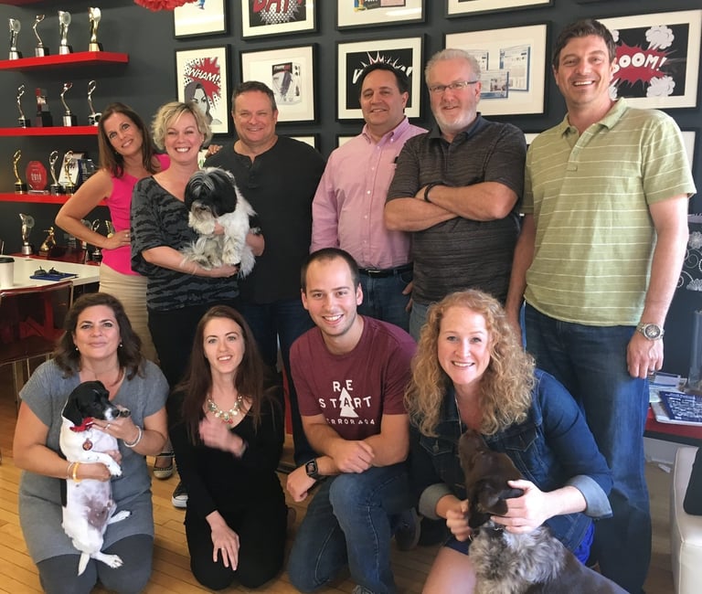Group of nine people posing together indoors, with four people holding small dogs, framed photos and awards visible on wall behind them