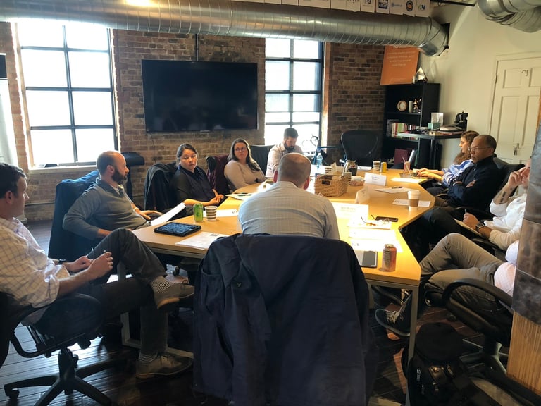 Group of eight people in business casual attire seated around a wooden conference table in a modern brick office with large windows and a TV screen on the wall