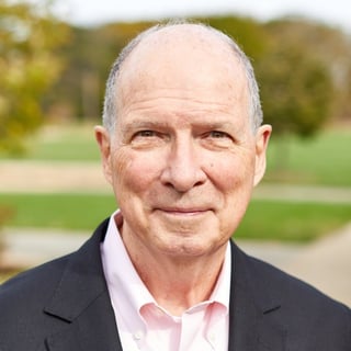 Portrait of an older man with white hair wearing a dark blazer and pink shirt, smiling outdoors