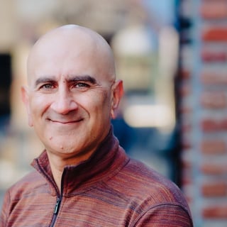 Bald man with a warm smile wearing a maroon zip-up shirt against a blurred urban background with brick buildings