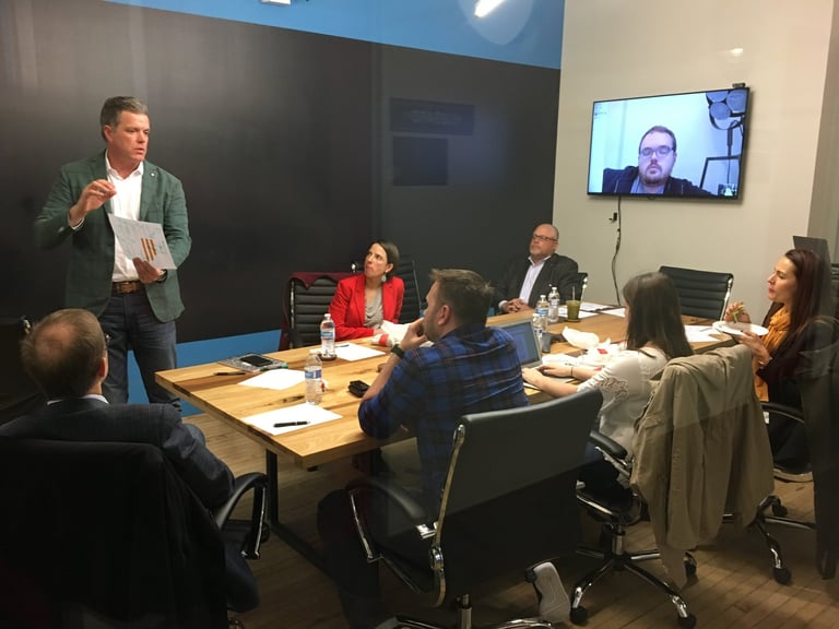 Business meeting with five people seated at a wooden table and one man standing, presenting to the group with a video call participant visible on screen
