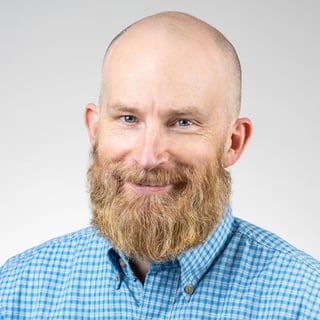 Bald man with full beard wearing blue checkered button-up shirt against gray background